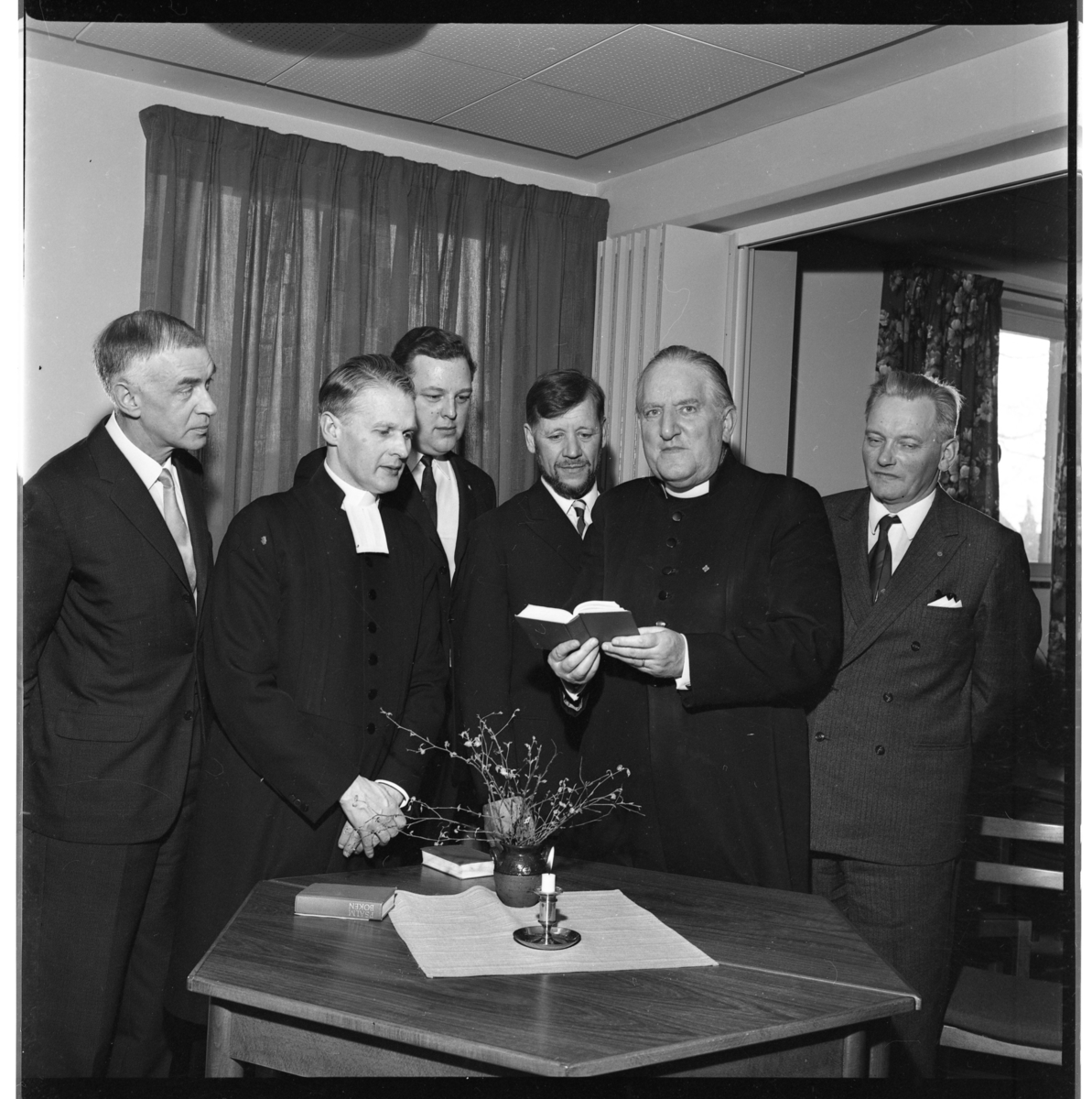Sex män varav två prästmän samlade runt ett bord med ett tänt ljus. Man tittar mot, troligen en psalmbok som kyrkoherde Wallis Wendel håller i händerna. De är från höger Göran Fong, Wallis Wendel samt Per-Axel Wenner.