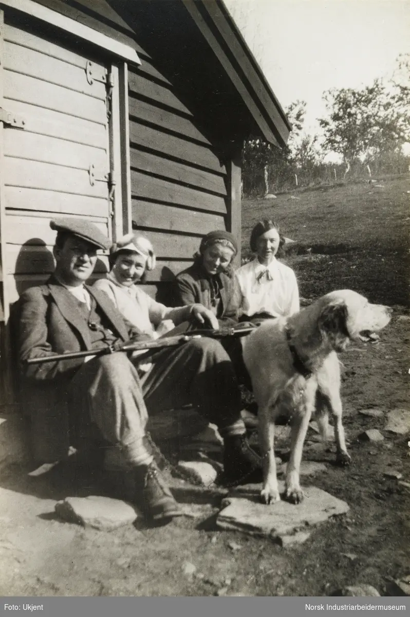 Familie og hund sitter langs hyttevegg. Mann med sikspens har gevær på fanget