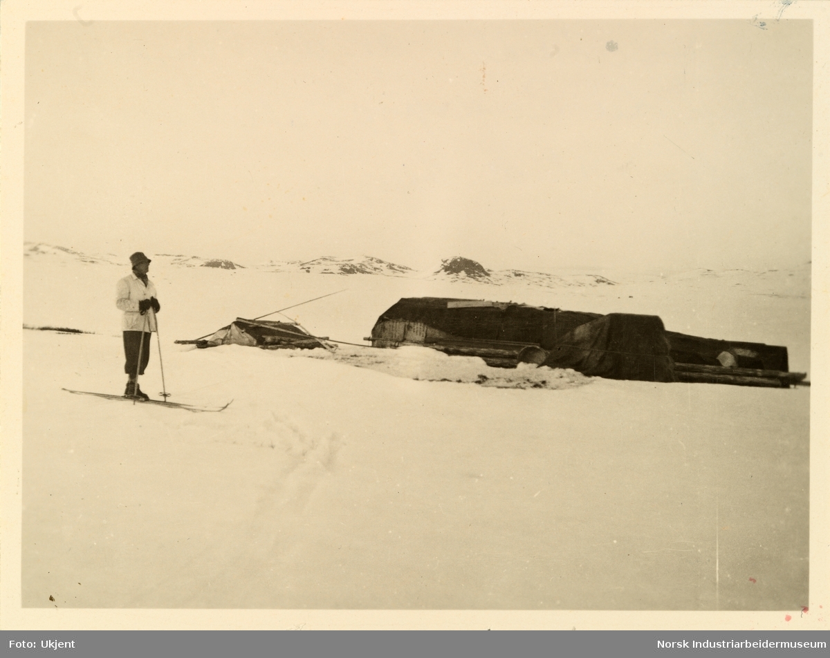 Mann i anorakk og med ski på bena står foran tildekt materiale til hytte i snødekt landskap