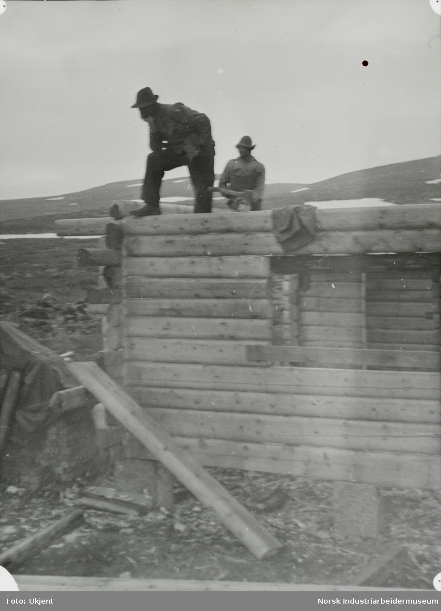 To menn står på laftet hytte under oppføring i fjellet