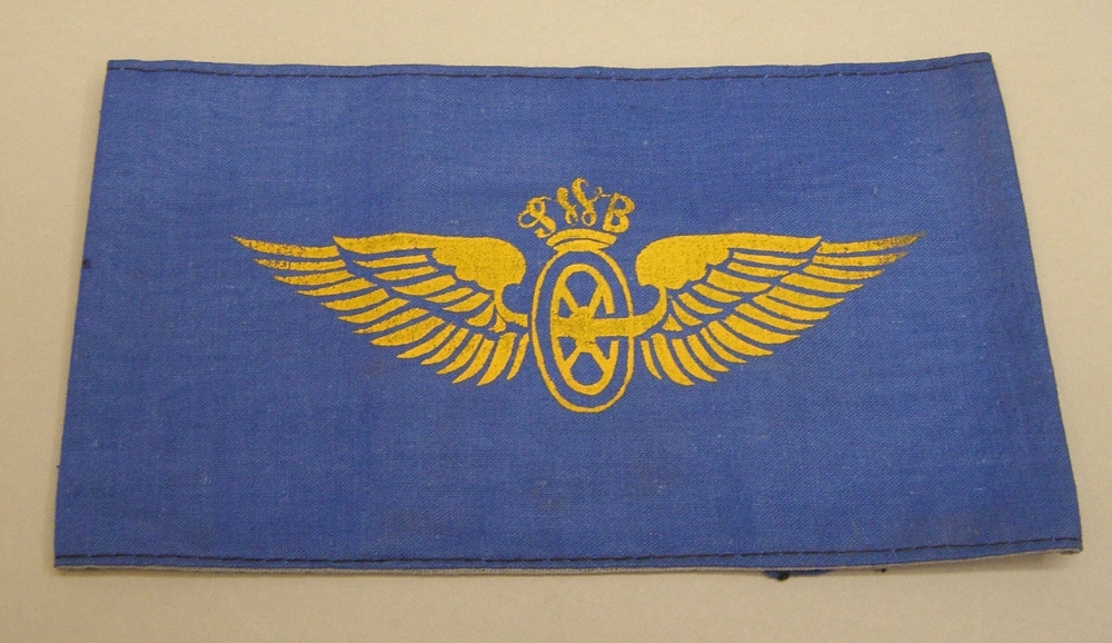 Armbindel av blått tyg med tryckt vinghjul i gult med SWB-monogram.