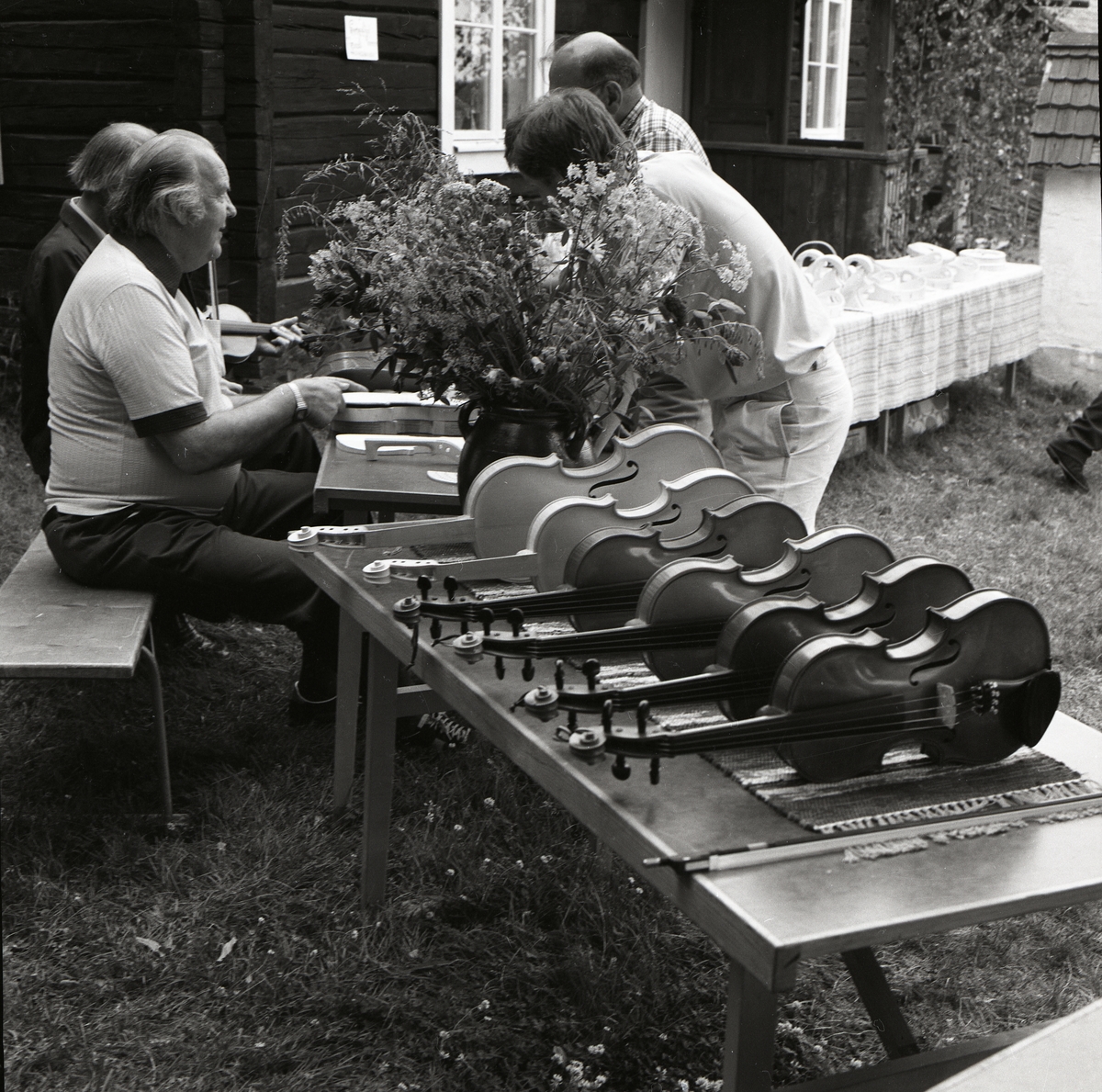 Fyra män samtalar vid ett bord. Bredvid dem står ytterligare ett bord där fiolinstrument står uppradade, Rengsjöfesten den 14 juli 1985.
