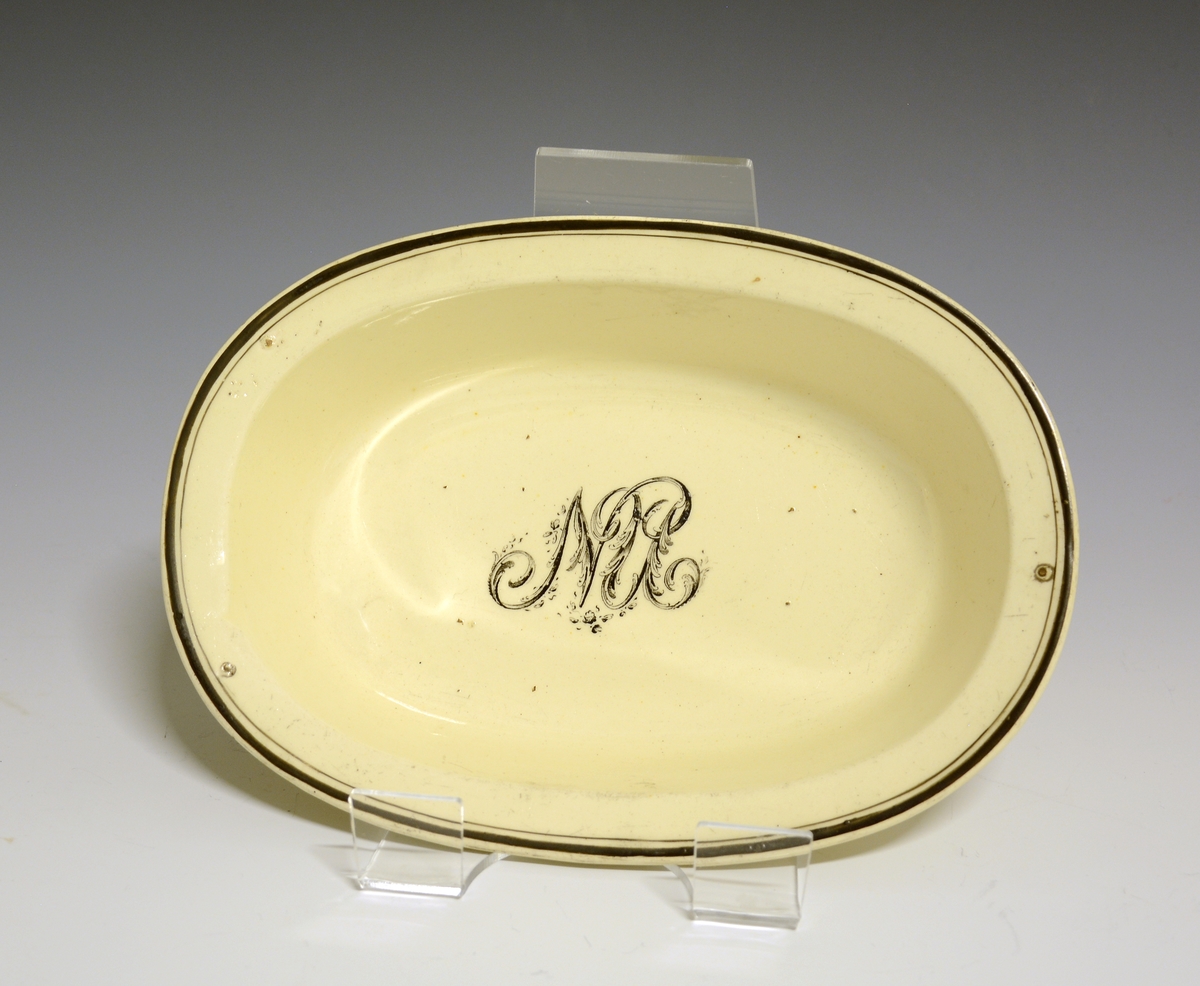 Lite, dypt, ovalt fat av stentøy med gulhvit glasur. I speilen et monogram med bokstavene NU. Ytterst på fanen en bredere og en smalere sort strek.