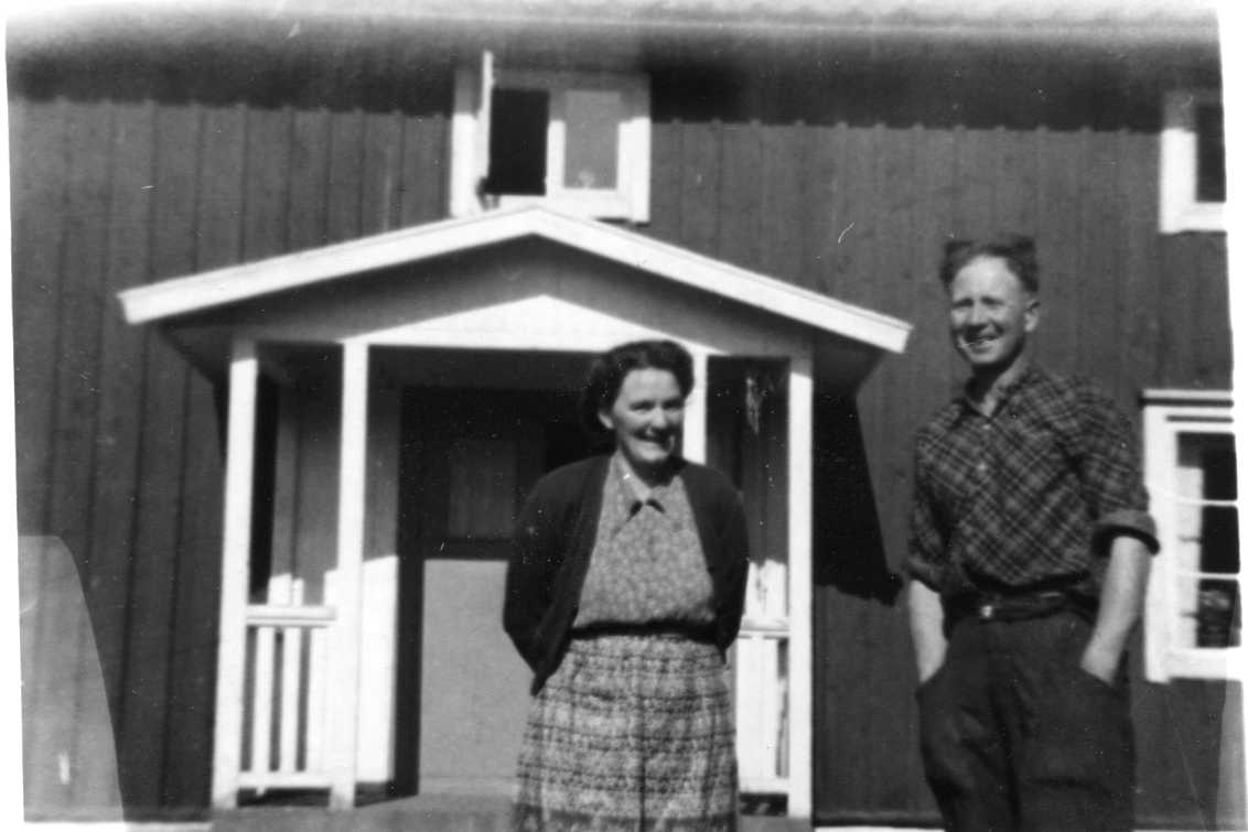 Märta Fransson med mönstrad blus, kofta och troligen förkläde står intill sin man Axel Fransson i rutig skjorta. De står framför verandan till sitt hus.