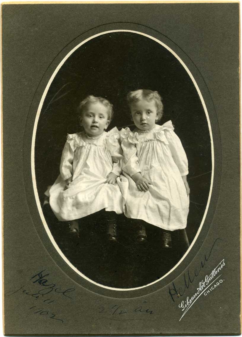Kabinettsfotografi: gruppbild med två små flickor i ljusa klänningar och kängor på fötterna, i en oval. Påskrift: "Hazel juli 11 1902 2½ år Hellen".