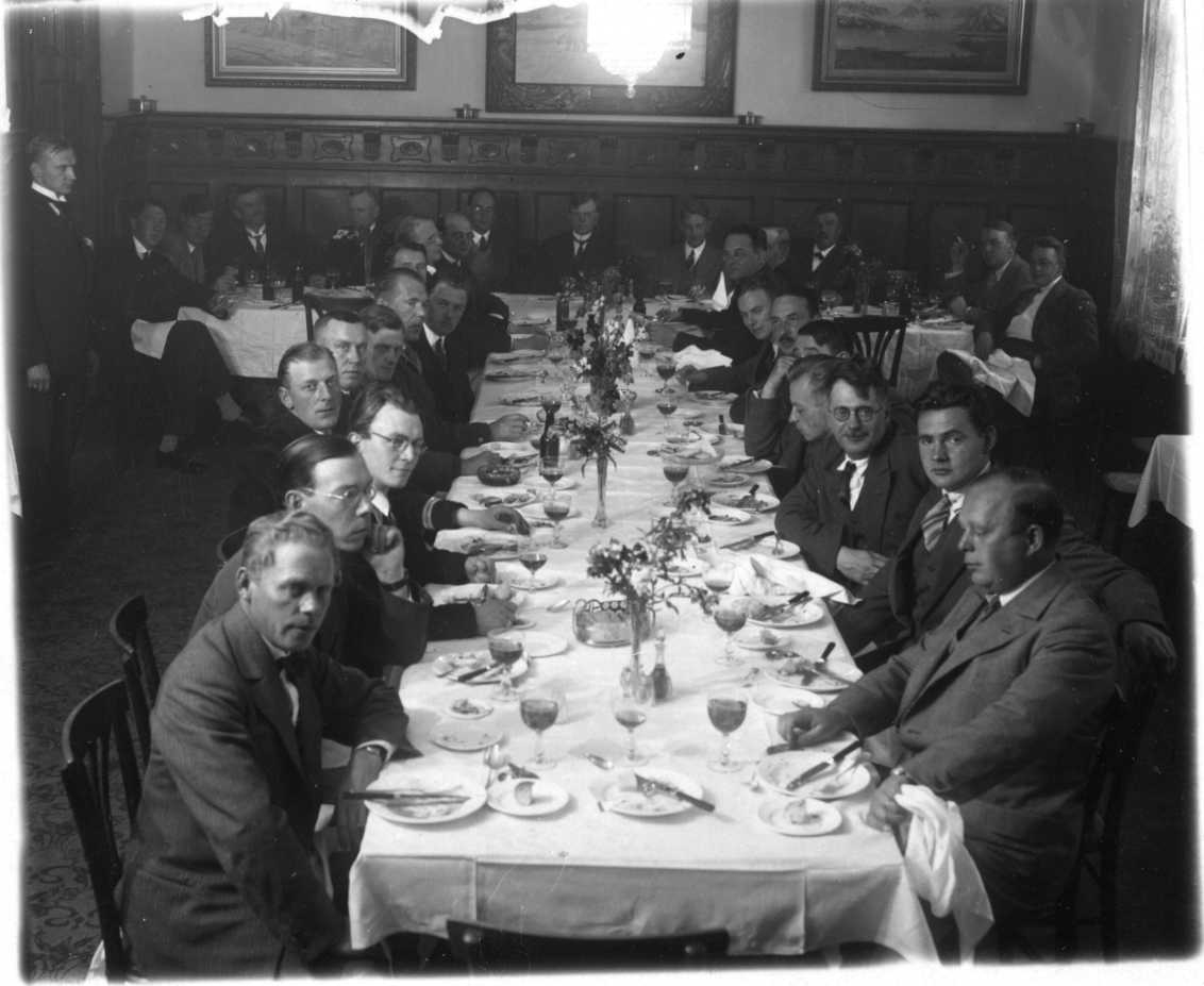 Ett 30-tal män sitter vid T-format dukat middagsbord med ljus duk. Maten är uppäten men man har dryck i glasen. Längst ner på vänstra sidan sitter sannolikt Sten Hedman. Till höger längst ner, som nummer tre från höger sitter sannolikt botanisten Olaf Hansen från fartyget Bratvaag.