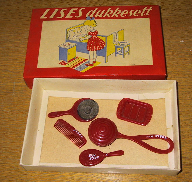 Lises dukkesett i en eske (a) som inneholder rangle (b), såpeskål (c), speil (d), børste (e), og kam (f) av rød plast.