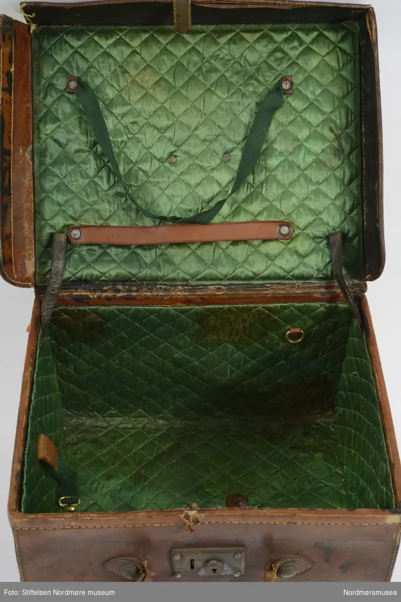 Koffert av skinn med messinglås med enkel lukkemekanisme i front. Innsiden er foret med grønn silke og lokket har to lærreimer som stopper den i å falle helt tilbake
