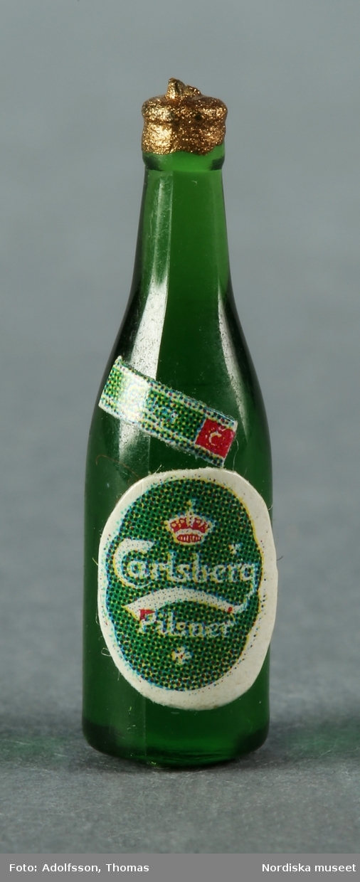 Två flaskor i miniatyr för måltidsdrycker, varav a) den bruna föreställer Coca-Cola och b) den gröna Carlsbergs öl. Båda är tillverkade av plast. Hör till inredningen i skafferiet bredvid frukostrummet på första våningen i dockskåp NM.331721+.