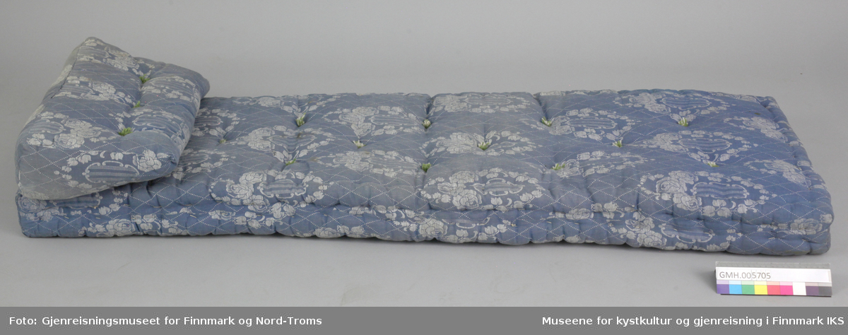Den blå madrass har en tilhørende kilepute. de er laget av tettvevd stoff med blå-hvit mønster. Fyllet holdes på plass av snøringer av naturhvite og grønne bomullstråder.  