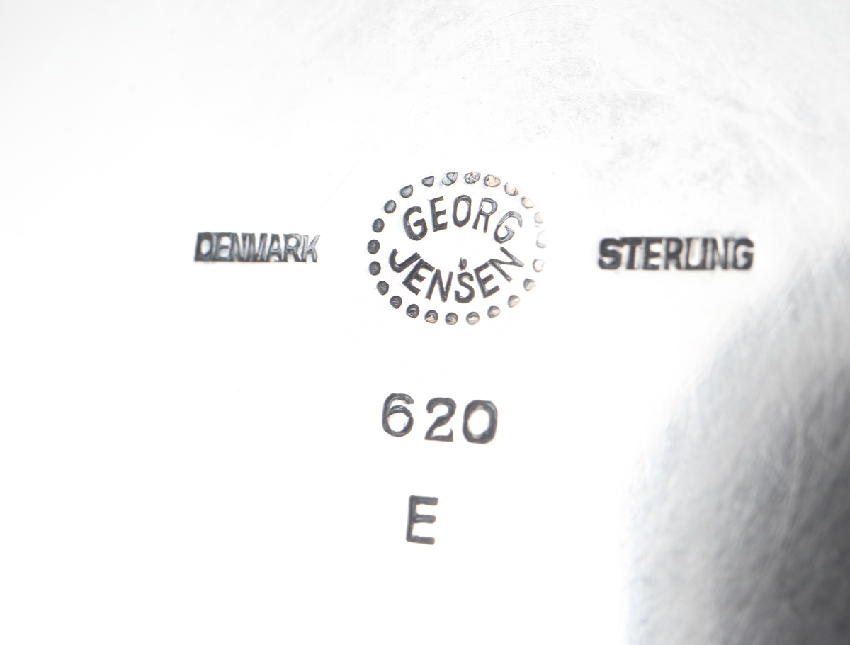 Fat av silver.
Rund, slät modell på låg, rund fot. På foten är graverat: "Fra Holmegaards Glasverk 8-9-1962". 
Inuti foten finns tillverkarstämpel m.m.

Inskrivet i huvudbok 1983.