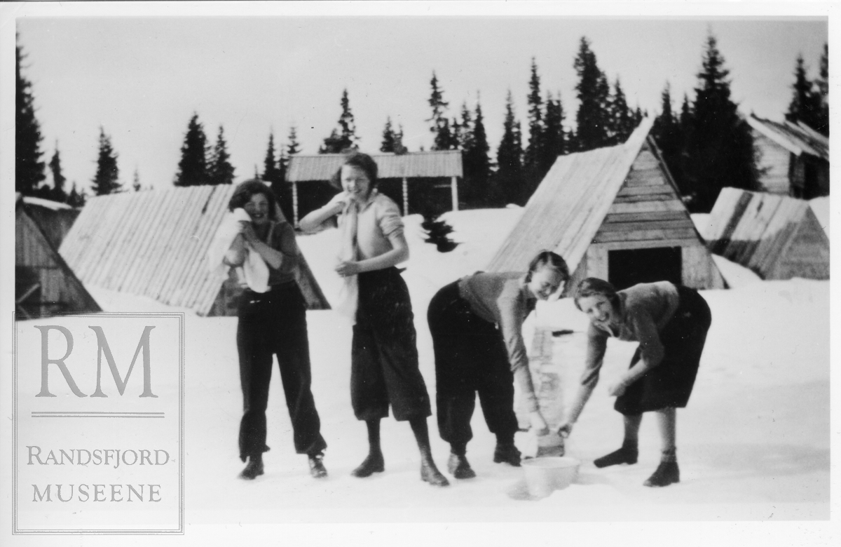 Fire jenter står på isen og vasker seg på Vassbråa. Båthus og hytter i bakgrunnen.