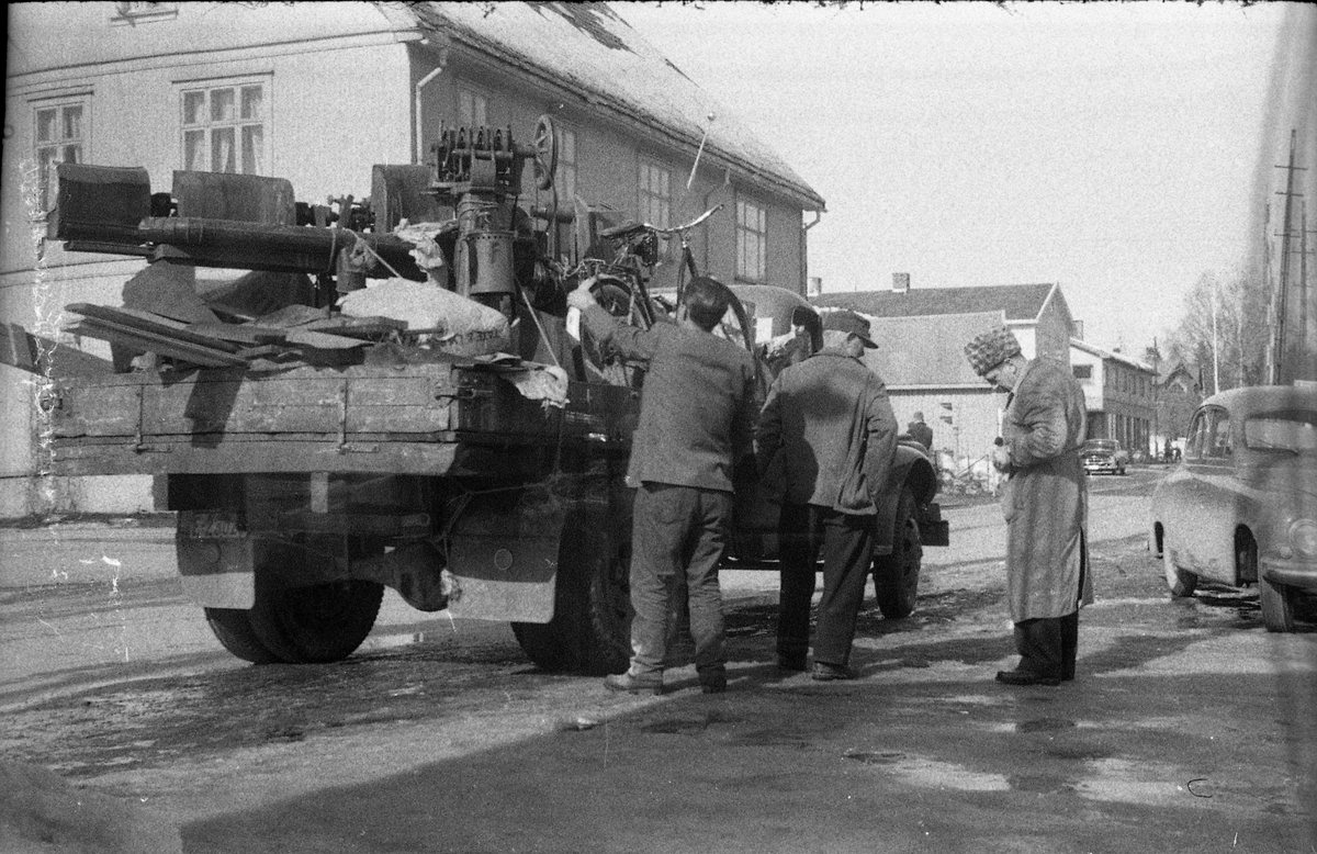 Gatebilde fra Lena april 1960. En lastebil lastet med skrot er parkert ved Brandt-Johannessens forretning. Mannen til høyre er ganske sikkert Arld Brandt-Johannessen, og de to ved lastebilen Harry Johansen (nærmest) og hans far Mauritz Johansen.