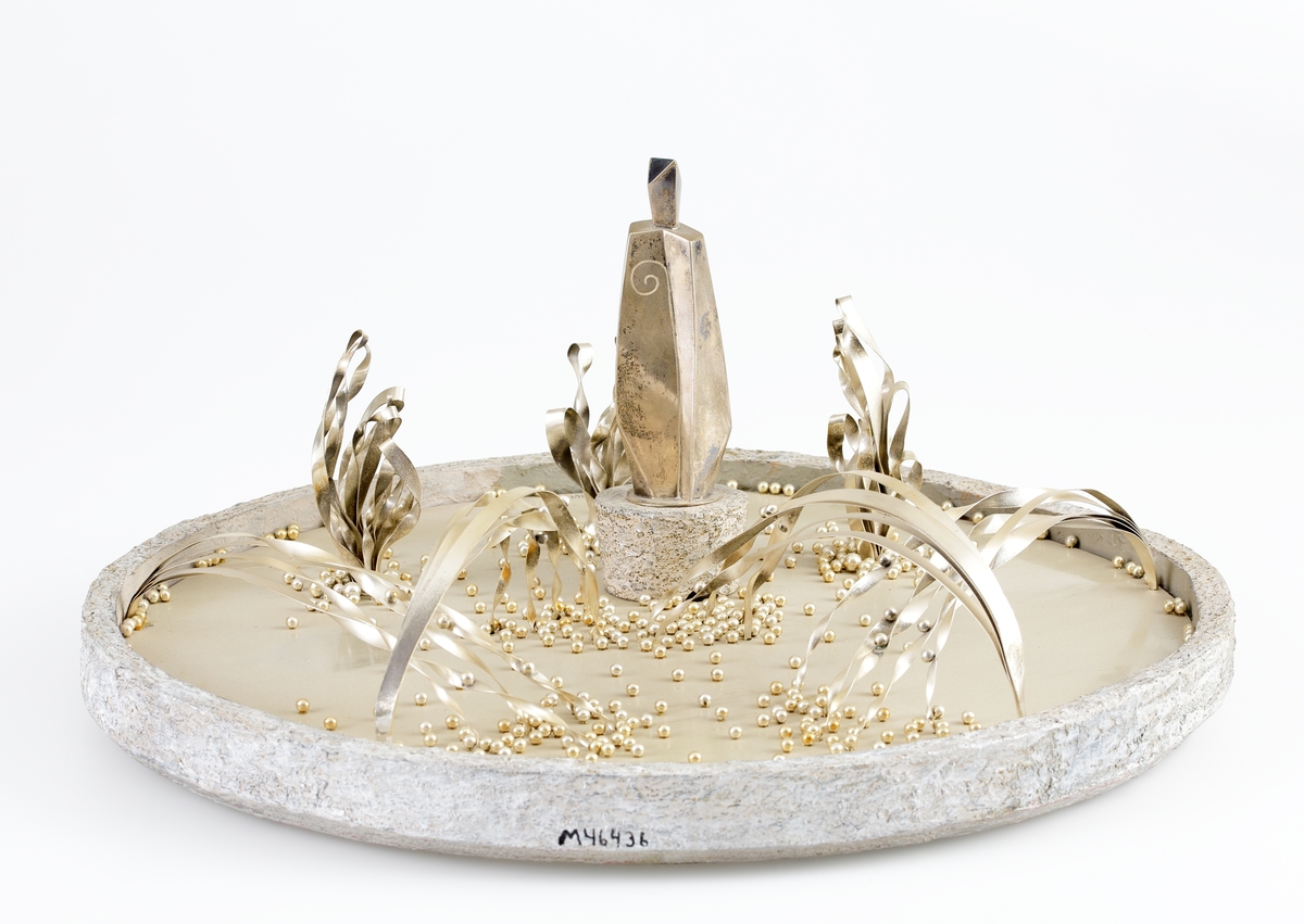 Modell i silver m.m. av Vicke Lindstrands skulpturgrupp "Legend i glas", (förslag) (Växjö).
Modellen har botten, sarg och S:t Sigfrids podium, tillverkat av färgad stenimiterad "Plastic Padding".
Vattenytan i modellen är tillverkad av en rund silverplåt, ca 44 cm i diameter. På ytan finns ett otal runda silverkulor, 
liknande droppar. Vid S:t Sigfrids sida finns "vattenkaskader", fyra knippen silverband med vridna ändar.
"Sunaman", ´"Unaman" och "Vinamans" skepnader framför S:t Sigfrid utgöres av tunna silverband vertikalt monterade i silverplattan, 
samt spiralvridna för att likna vattenkaskader.
S:t Sigfrids figur är tillverkad av drivet silver, 14,7 cm hög med biskopskräklan ingraverad på framsidan. Figuren är fastlödd på 
en cirkelrund silverplatta som är försedd med stämplar m.m.
