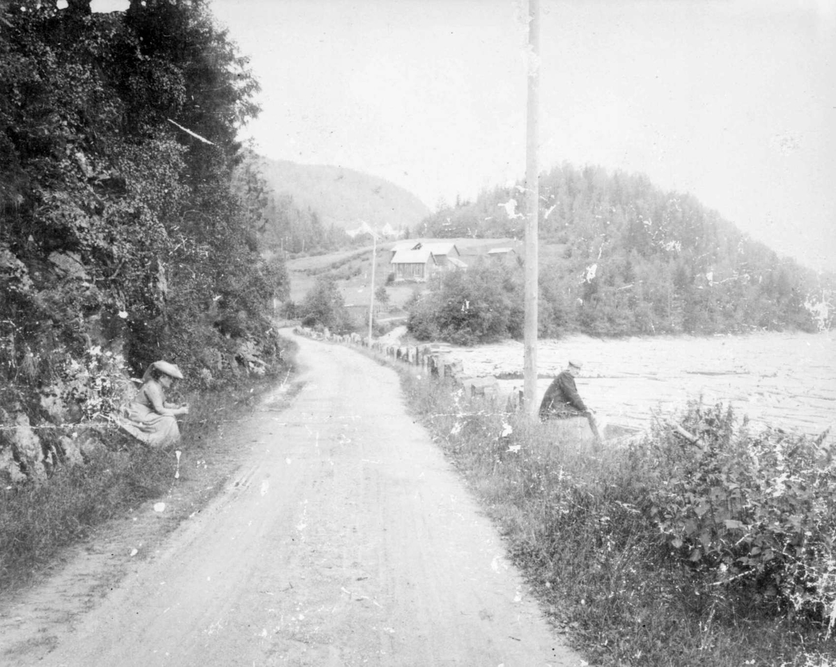 Repro: Jørstadmovegen i Leirvika rett sør for krysset med Kastrudvegen, en kvinne og en mann sittende ved veien, gårdsbruk/småbruk i bakgrunnen.