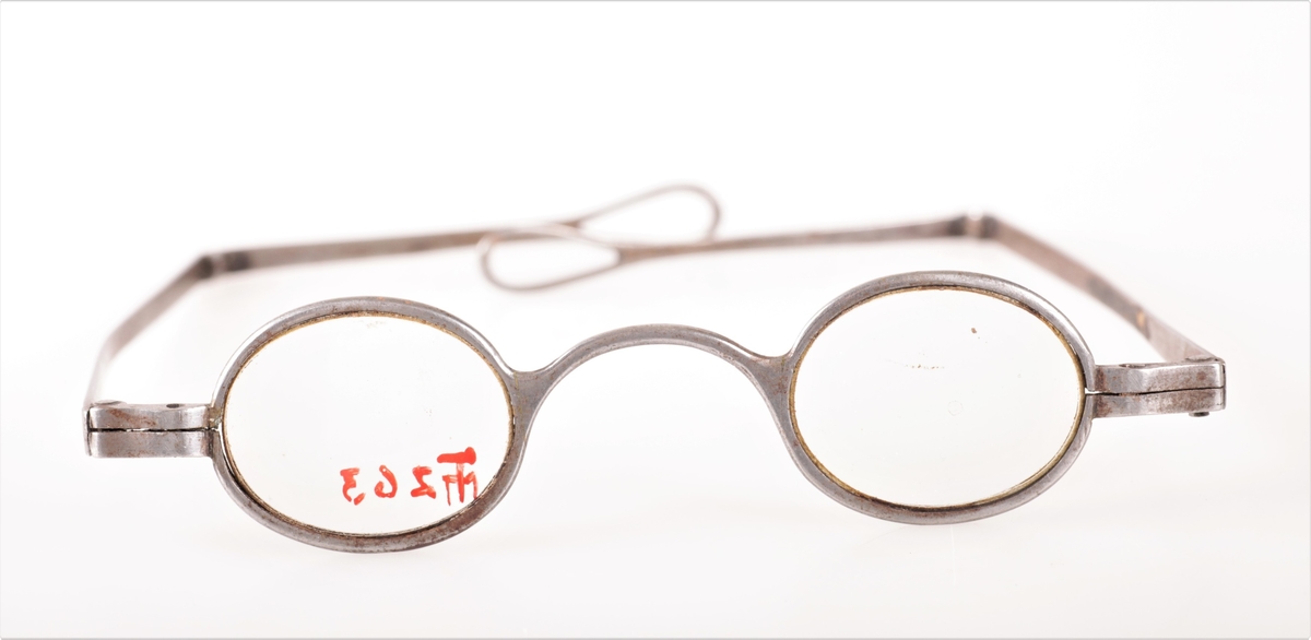 Ovale brilleglass i stålinnfatning, flate stenger med ledd som bøyer inn. Ytterst på leddet stort pæreformet hull