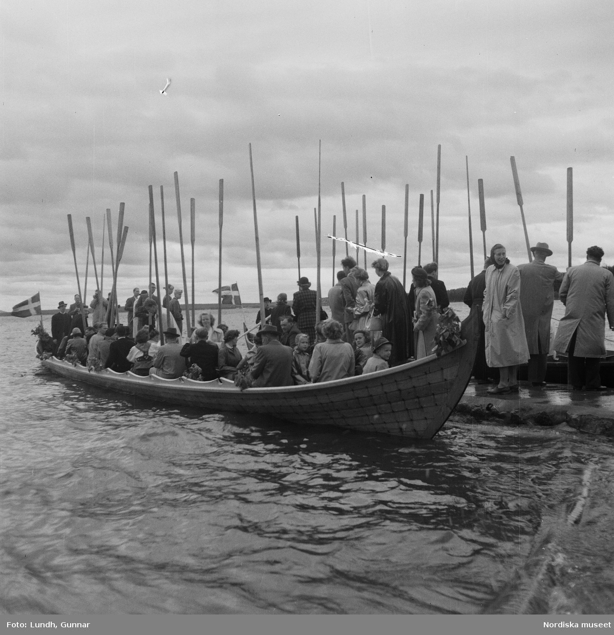 Motiv: (ingen anteckning) ;
En folksamling står vid en brygga med två kyrkbåtar, kvinnor och män ror kyrkbåtar i samband med midsommarfirande.