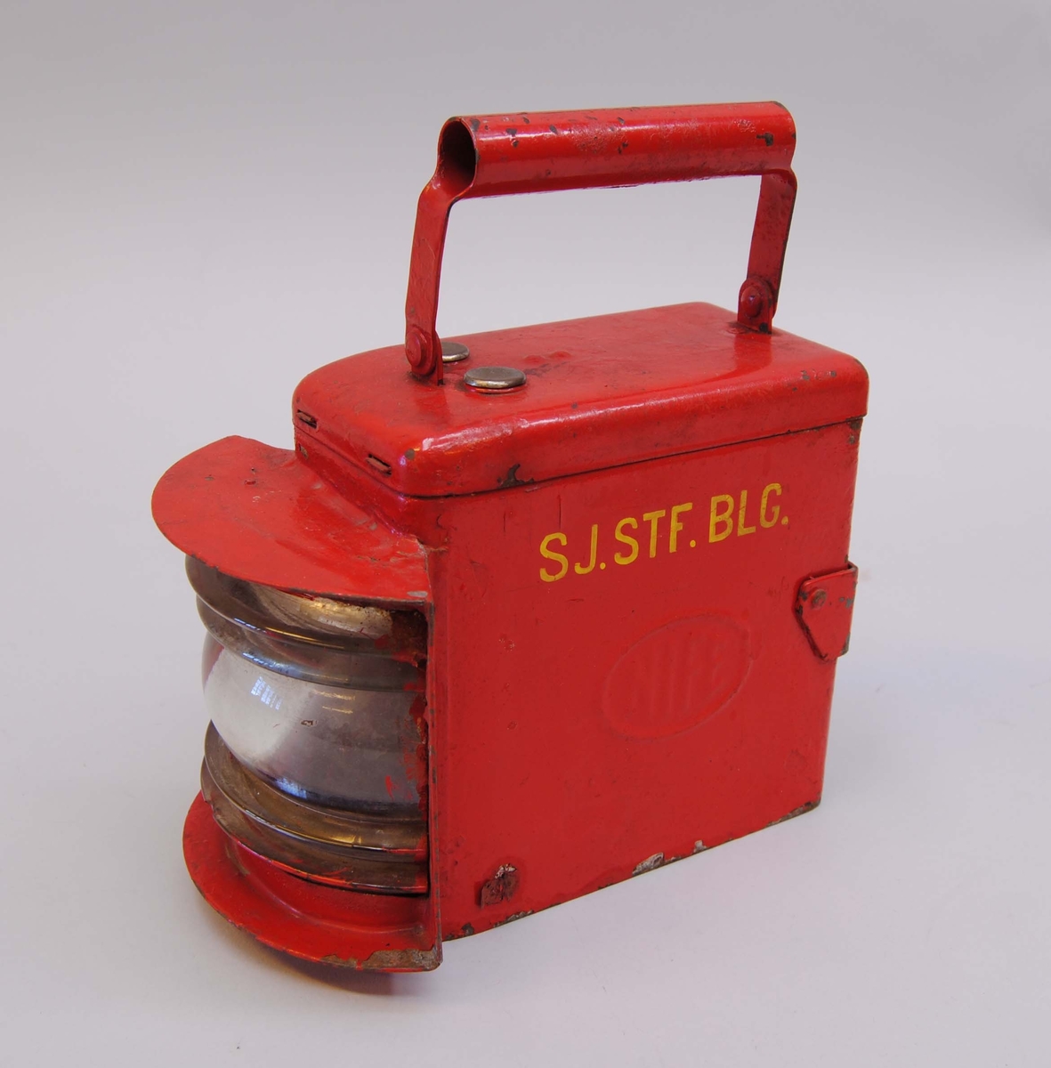 Röd batteridriven signallykta märkt "SJ. STF. BLG." med gult. På sidorna står det "NIFE" i plåten. På ovansidan finns ett handtag och på baksidan en hållare.