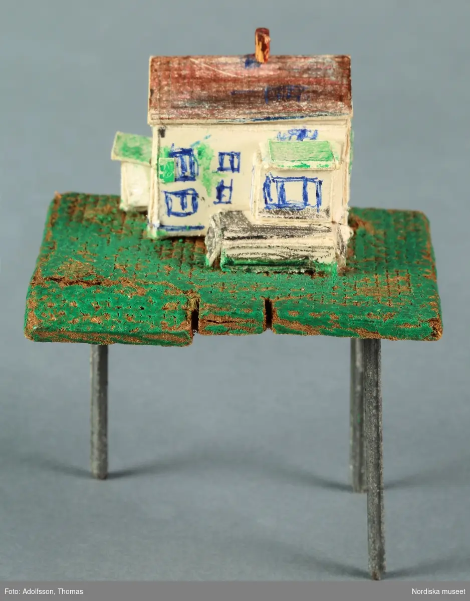 En hemtillverkad miniatyrmodell av dockskåpet NM.0331721+. Modellen av 1930-talsvillan är gjord av pappkartong och står på en grönmålad platta av masonit. Plattan är försedd med tre bordsben av metallspikar. Ett ben saknas. Påmålade fönster och påklistrade fönsterluckor. Hör till inredningen i hobbyrummet i dockskåpets källare.