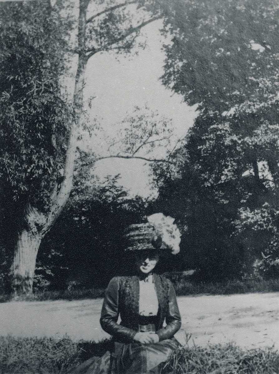 Ung kvinne i ført hatt med pynt og drakt med langt skjørt sittende i en park eller hage. 