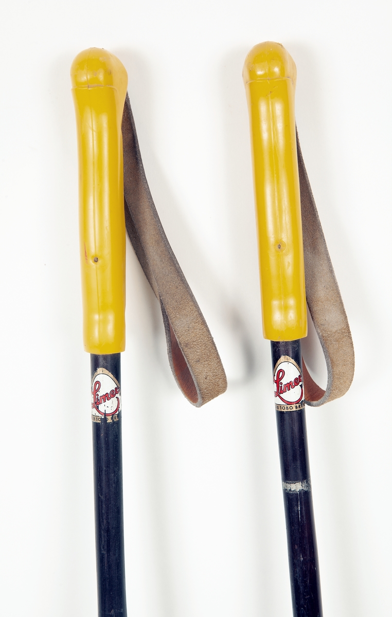 Två skidstavar i bambu, målad med brun/svart färg. Skinnholk av lättmetall och järndubb, rottingkringla med korslagda läderremmar fästa med skinnrem. Greppet i gul plast, handrem av läder.