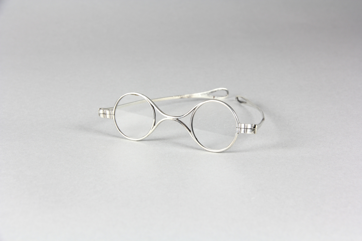 Glasögon, mindre glas och smala bågar av silver.  Två skalmar som utåt avslutas med droppformade öglor.