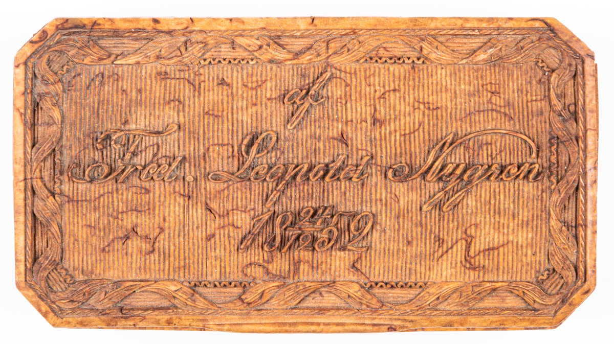 Snusdosa med fällock, av masur, avlång med rikt utskuren ornering.
Märkt: "af Fredr. Leopold Nygren 24/12 1852.