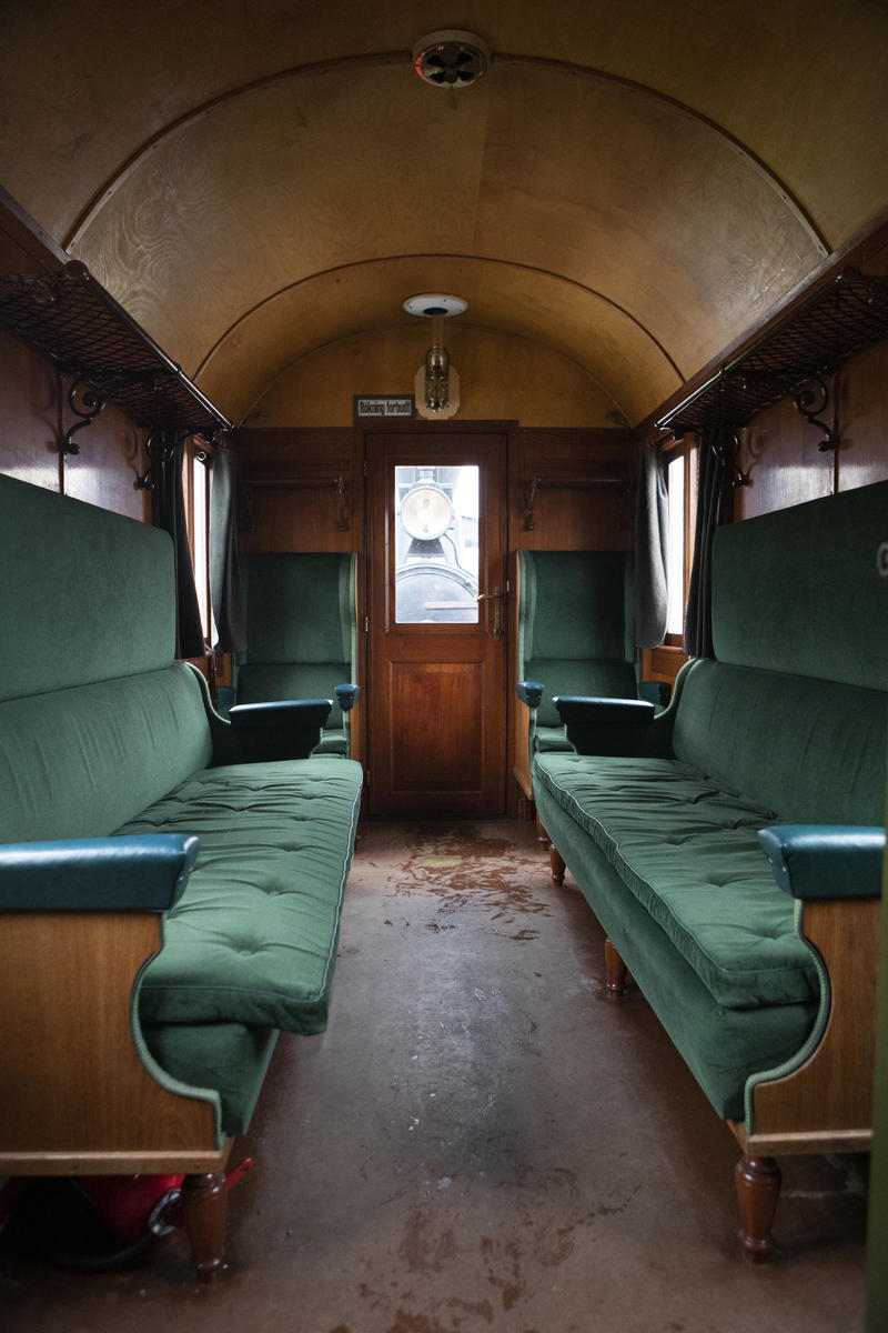 Interiør fra en andre-klasses passasjervogn på Urskog-Hølandsbanen. To tre-seters sofaer i flaskegrønn plysj er plassert mot hverandre i vognens lengderetning. To stoler har ryggen mot kortveggen, en på hver side av utgangsdøren.