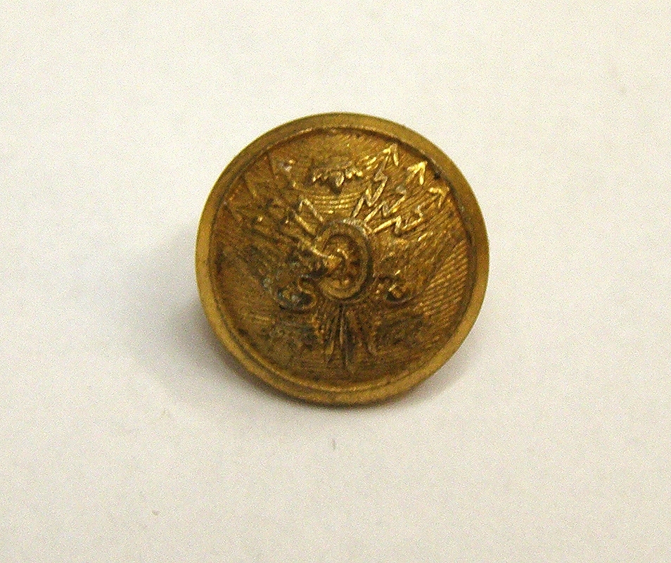 Mösstränsknapp av förgylld metall, med gyllene vighjul och tre kronor på räfflad botten. Knappen är obottnad.