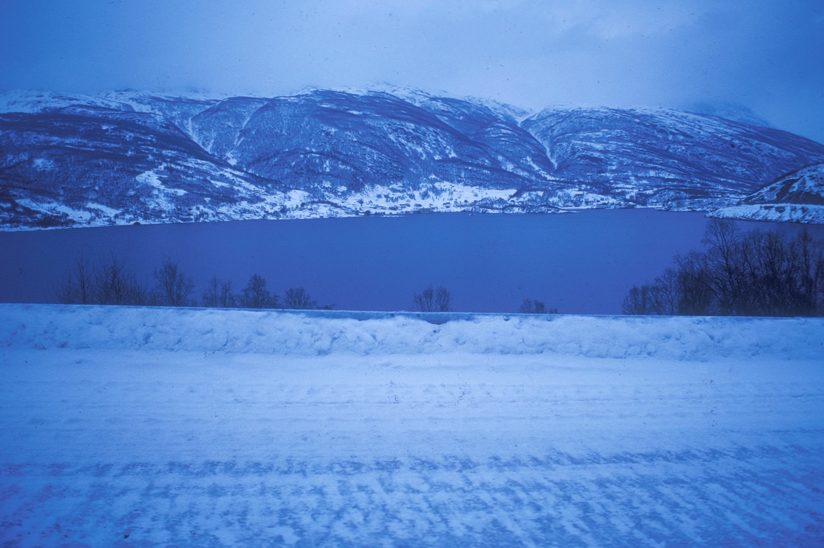 Snøkledd kystlandskap. En fjord omgitt av fjell. Bildet er tatt fra veien, som ses i forgrunnen.