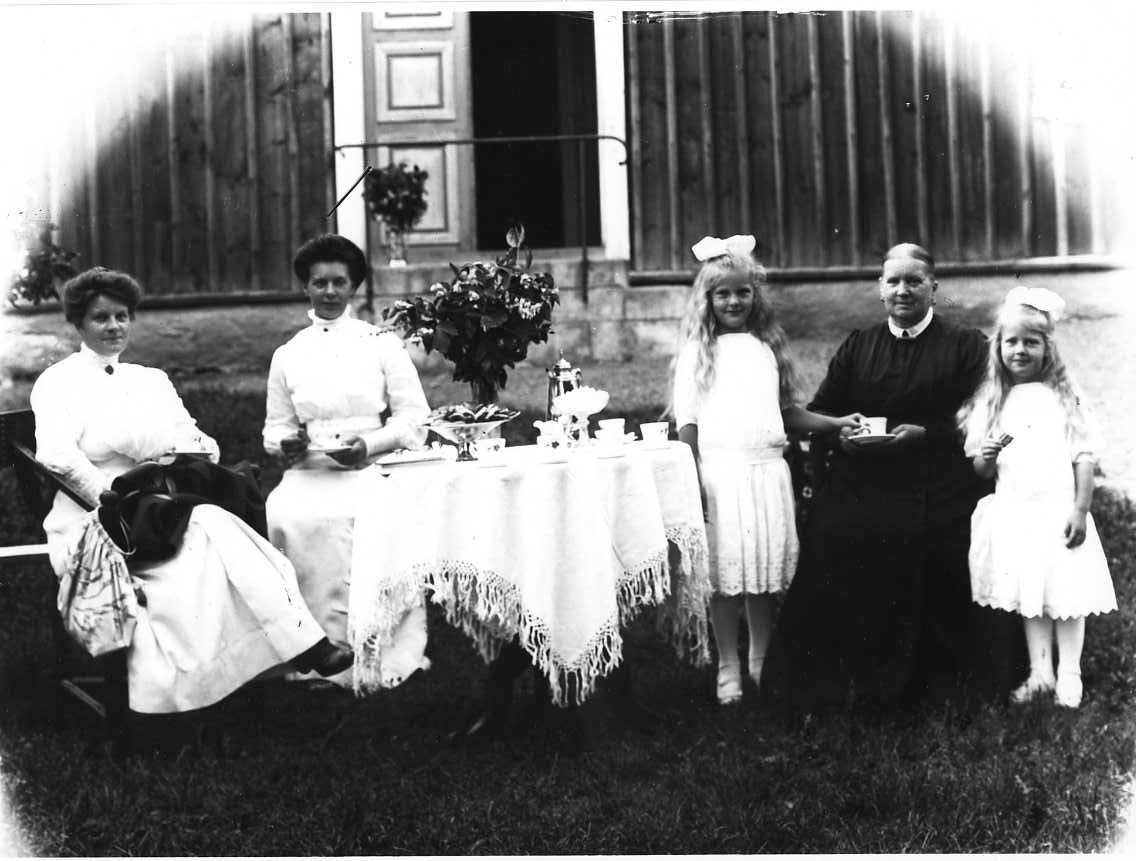 Gruppbild med tre kvinnor och två flickor som sitter och står vid ett dukat kaffebord utomhus framför Missionshuset i Örserum, en rödfärgad byggnad med locklistpanel. Till vänster sitter två kvinnor i ljusa klänningar, det är syskonen Agnes och Tyra Josefsson. Till höger två flickor i ljusa klänningar, långt hår med rosett i håret flankerar en äldre kvinna i mörk klänning som håller en kaffekopp i knät. Flickorna är Agnes döttrar och den äldre kvinnan är mormor Sofie Josefsson.