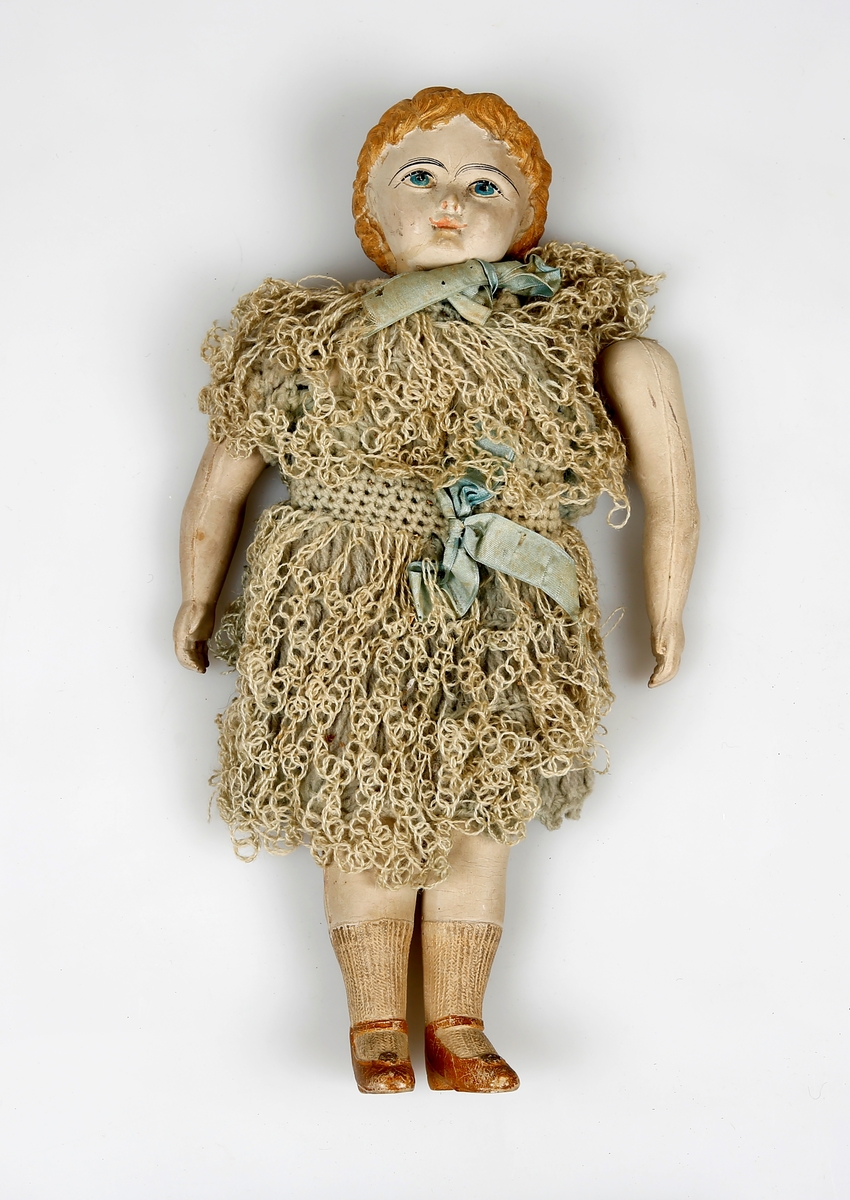 Dukke med plastkropp og -hode. Hode og armer er festet til resten av kroppen. En arm er løs. Dukken har heklet kjole. 