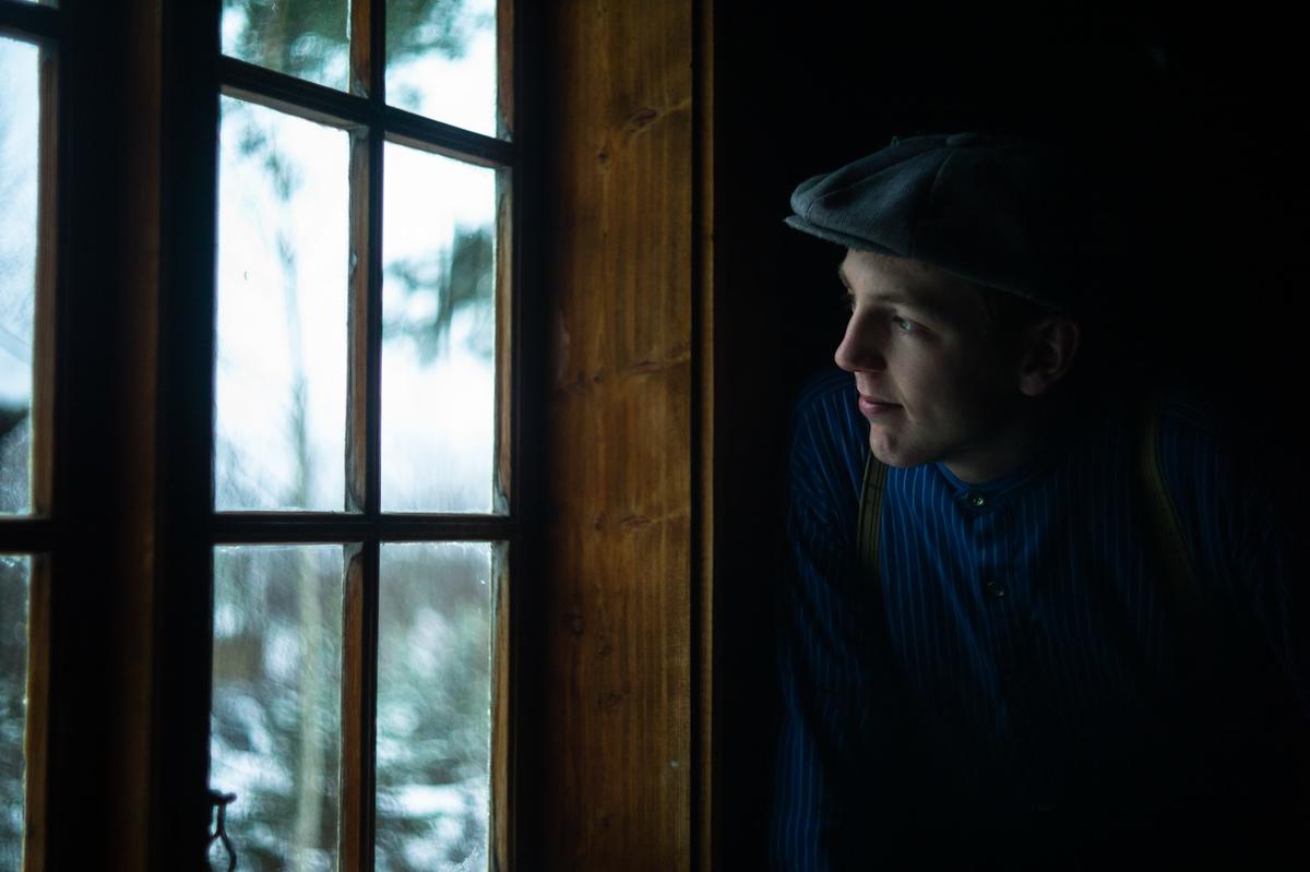 Fotografiet er tatt under arrangementet Jul i stuene. En ung gutt titter ut av et vindu. Ute er det lyst, og det er snø på trærne. Gutten har på seg busserull og lue.