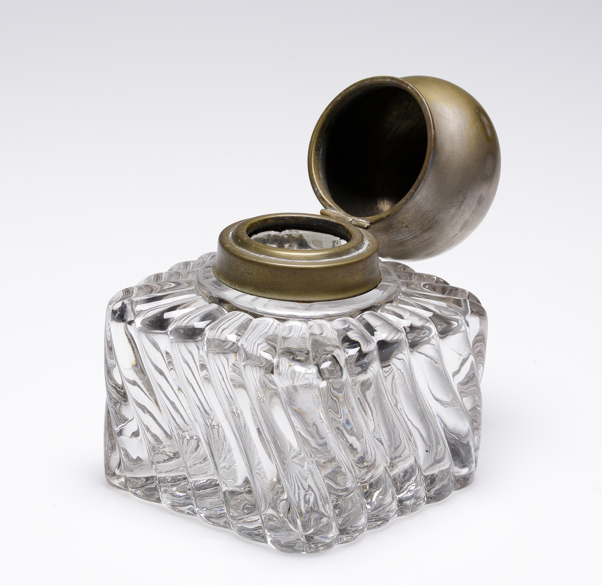 Bläckhorn i pressat glas med fyrkantig form och snedgående räfflor. Lock av mässing. Tillverkare är troligen Baccarat.