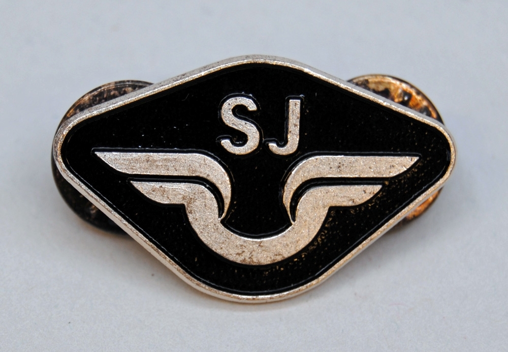 Metallbricka med två nålar på baksidan för kunna sätta fast på uniformen. På brickan finns SJ:s nya logotyp från 1991 och över den står det "SJ", bakgrunden är svart medans logotypen och texten är metallfärgad. På baksidan är det ett litet tryck med texten "Sporrong".