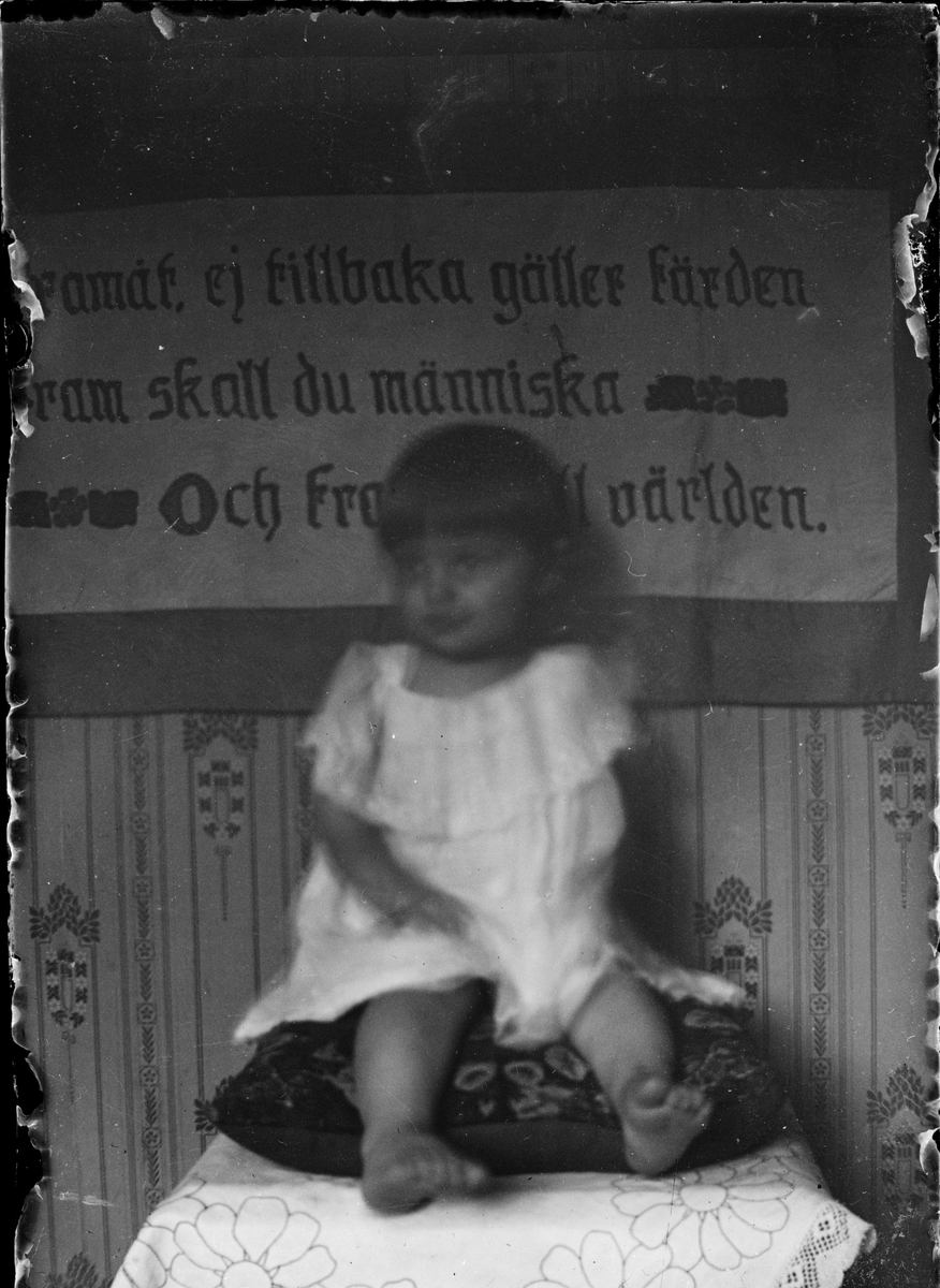Ett barn sitter på en kudde med en broderad bonad bakom sig, Östhammar, Uppland