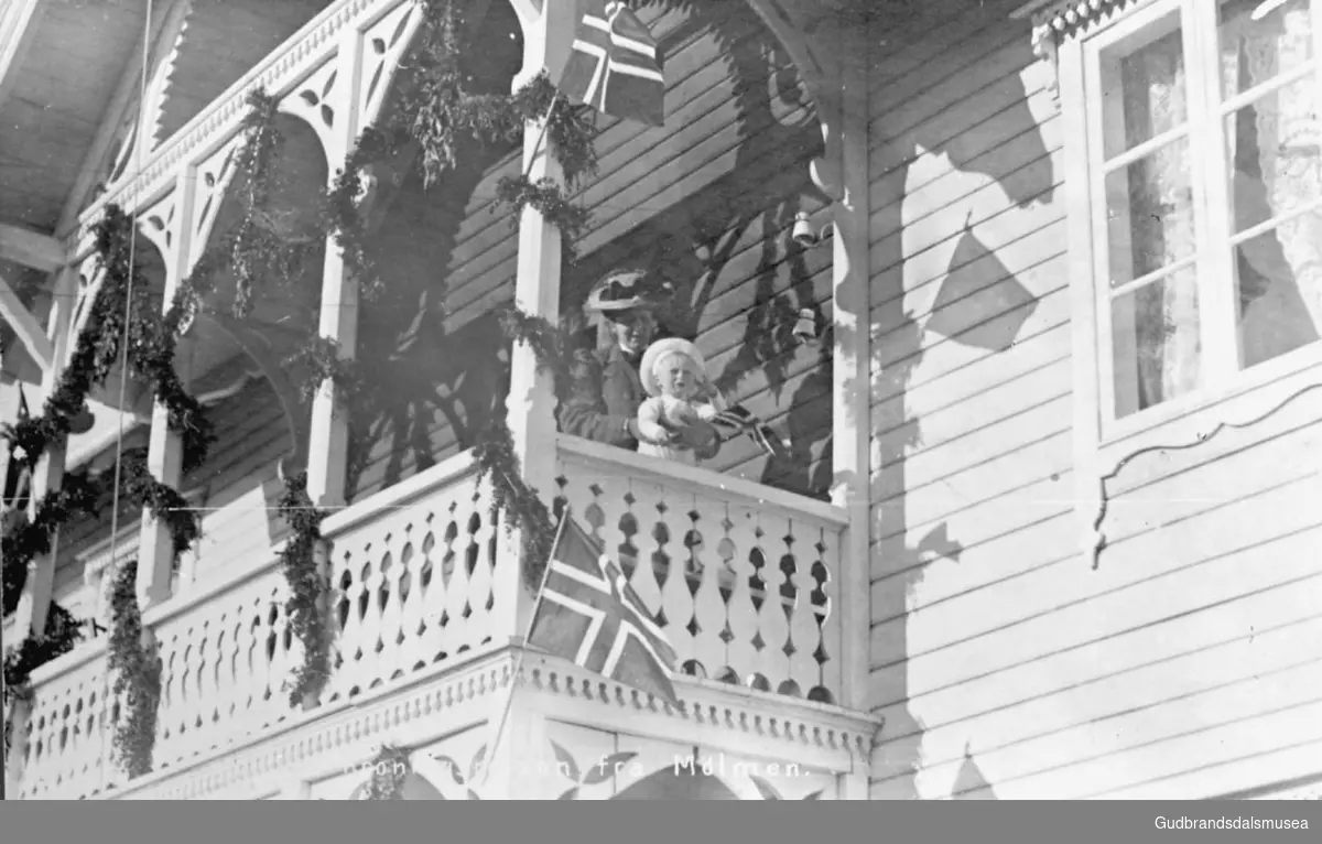 Fra kroningsreisa i Mølme, i 1906. Motiv av en kvinne på balkongen, hun holder et barn, stor toetasjers bygning pyntet utvendig ved balkong og inngangsparti, det norske flagg henger oppe.