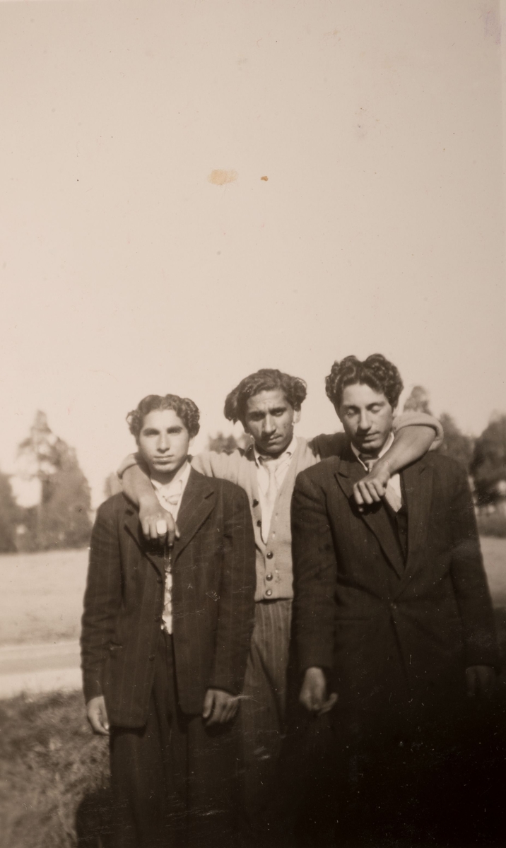 Tre romska män i ett läger i Sandviken, juni 1947. Mannen i mitten har lagt armarna om de två männen som står på var sin sida om honom.