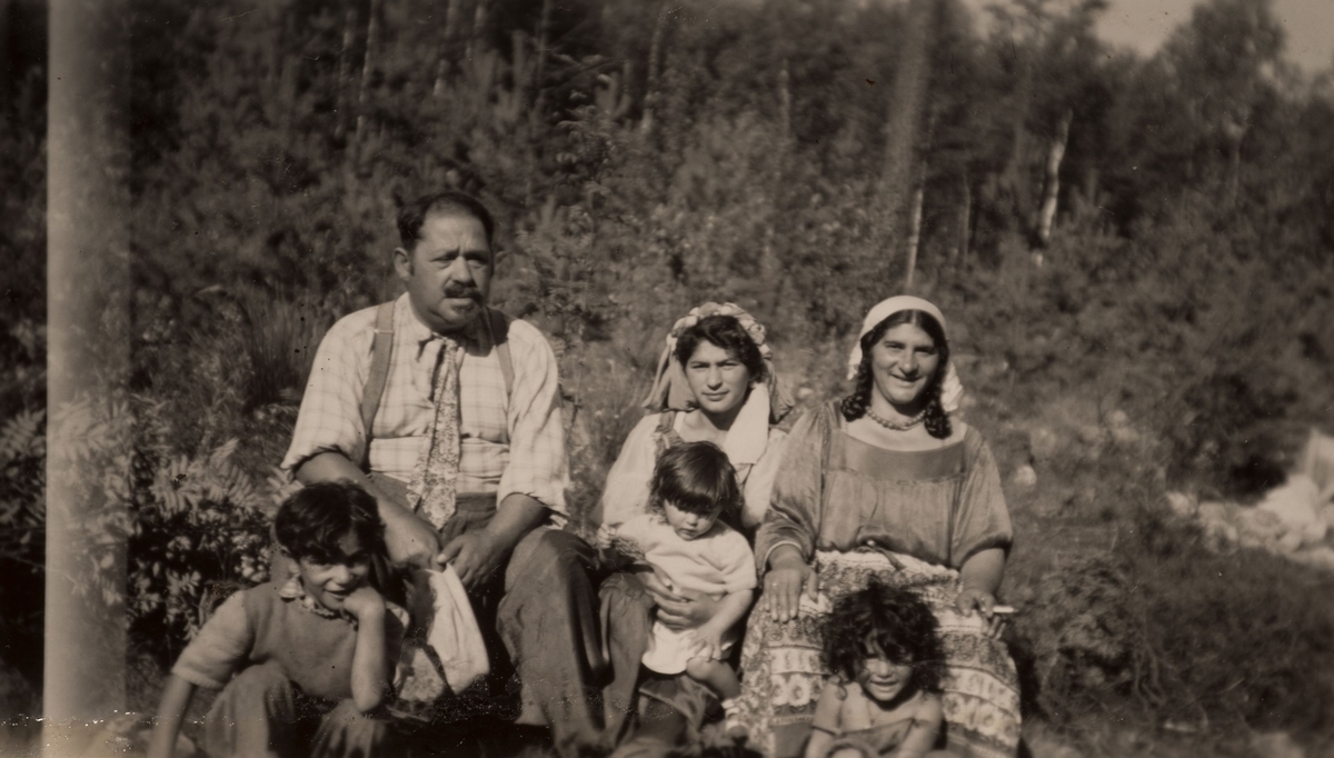 En grupp romer samlade för fotografering, en sommardag i Hofors 1950. I mitten sitter en yngre kvinna med ett barn i famnen, flankerad av en äldre man och en kvinna med varsitt barn vid fötterna.