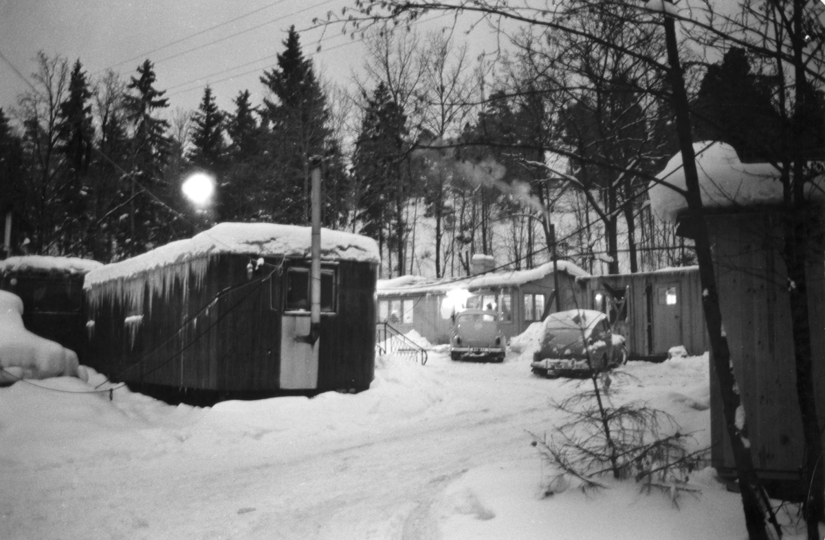 Romskt läger i Ekstubben 1963. Lägerplatsen låg i Skarpnäck söder om Stockholm, i nuvarande Flatens naturområde, på en plats där det i dag ligger en motorbana. Lägret var samtida med Skarpnäckslägret några kilometer bort och iordningställdes 1959 av Stockholms stad för svenska romer i kommunen som ännu inte hade fast bostad. Ekstubben stod färdig för inflyttning 15 mars 1960. För drygt 50 år sedan var Ekstubben och Skarpnäckslägret i fokus för Katarina Taikons kamp för att romernas rättigheter, men i dag är det få personer som känner till dem och vet var lägren legat.

Bilden är tagen i samband med reportaget ”Vagabond eller vanlig människa?” av Roland Hjelte och Karl Axel Sjöblom som sändes i SVT i mars 1963. Reportaget skildrar livet i lägren i Ekstubben och Skarpnäck under vintern 1963. I intervjuerna med de boende i lägret framhålls det akuta önskemålet om fast bostad. I lägret bodde det året 15-20 familjer och reportern berättar att människor har bott här flera år i väntan på att Stockholms stad ska lösa bostadsproblemet. Det är vinter, snön har fallit och vissa nätter går temperaturen under nollstrecket inne i husvagnarna. En man berättar att han är uppe hela nätterna för att elda i kaminen. Han säger till reportern: ”När alla andra fått bekvämligheter ska vi också ha det. Vi kan inte ha det så här!”.

Fastighetskontoret i Stockholms stad hade i en kommunal utredning 1955–1957 kommit fram till förslaget att iordningställa särskilda lägerplatser. Lägren skulle finnas under en övergångsperiod innan fasta bostäder ordnats, men i realiteten drog bostadsfrågan ut på tiden och lägren blev kvar i fem år. I Ekstubben bodde människor under enkla förhållanden, inledningsvis i tält och vagnar och efter hand också i egenbyggda baracker och stugor kring en öppen yta. Lägret försågs med el och vatten, torrtoaletter samt sophämtning. 

Det skulle dröja till maj 1964 innan de sista av de svenska romerna flyttade från Ekstubben. Senare användes Ekstubben dock fortsatt som lägerplats för romer som kommit till Sverige från andra delar av Europa.