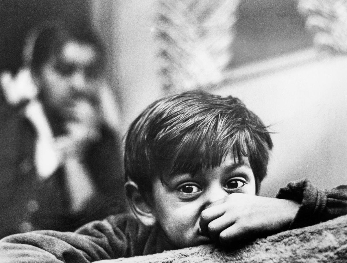 En romsk pojke tittar in i kameran. Han lutar sig mot något och hans ena hand döljer delvis hans ansikte, I bakgrunden syns en romsk flicka.