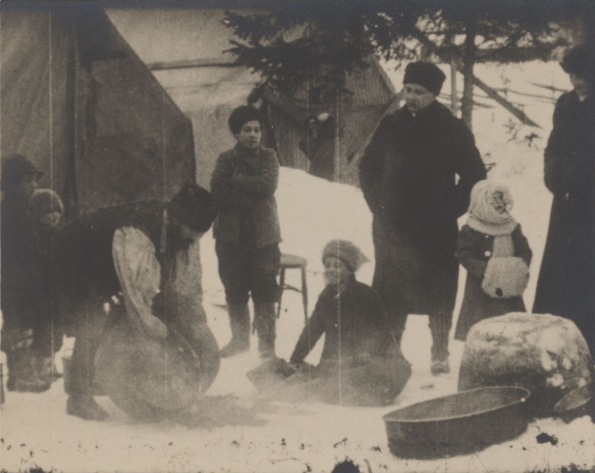 Människor i olika åldrar har samlats kring en eld i snön. Mannen till vänster ägnar sig troligen åt förtenning av ett kopparkärl. Personerna på bilden är Kelderashromer och kom till Sverige via Ryssland och Finland.