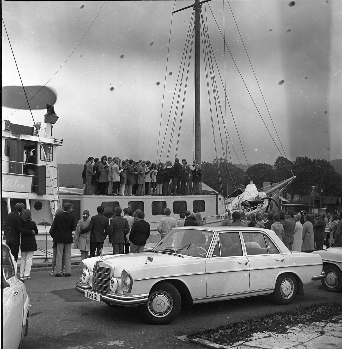 Missionsbåten Elida ligger vid kajen i Gränna hamn. En kör på båten sjunger för publik på kajen. En Mercedes Benz i förgrunden.