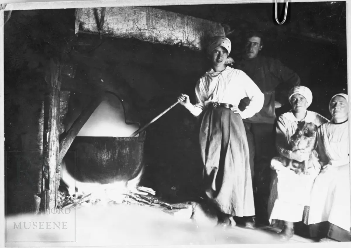 Ei jente står og rører i en stor gryte som henger over åpen ild i en peis. To kvinner sitter ved siden av og en mann står bak dem.