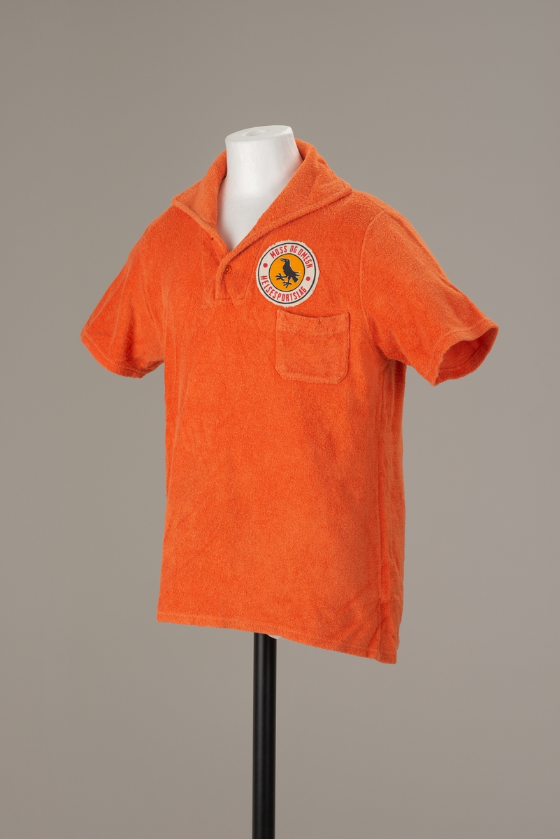 Oransje pique skjorte med krage med to knapper. Korte ermer. Brystlomme på venstre side.