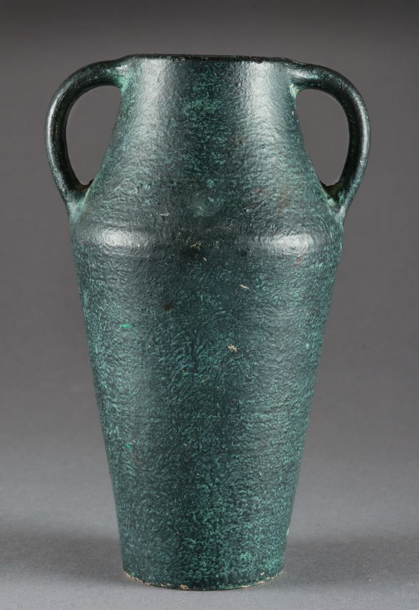 Vas med två hänklar av lergods. Handdrejad och glaserad och målad i s.k. grön patinering.
Vasen är omärkt men förmodligen från Bo Fajans i Gävle.