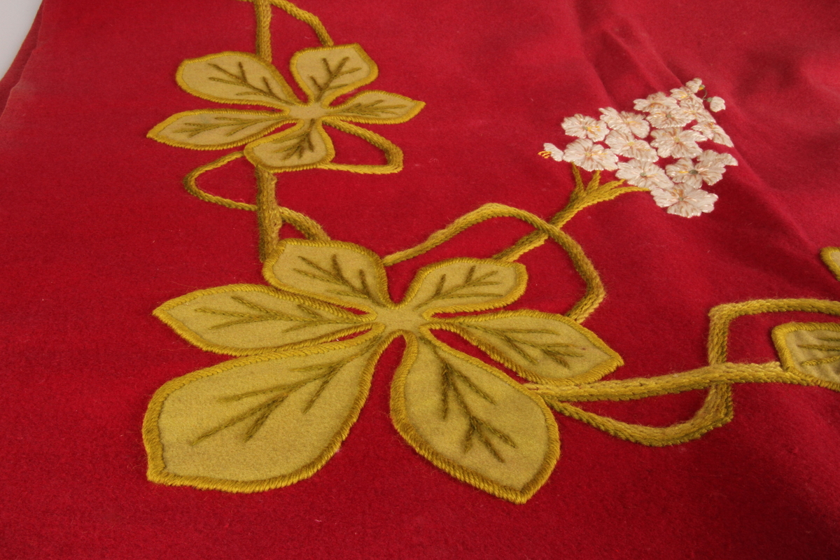 Duk brodert med blomst- og bladmotiv, i grønt og hvitt på rød bunn.