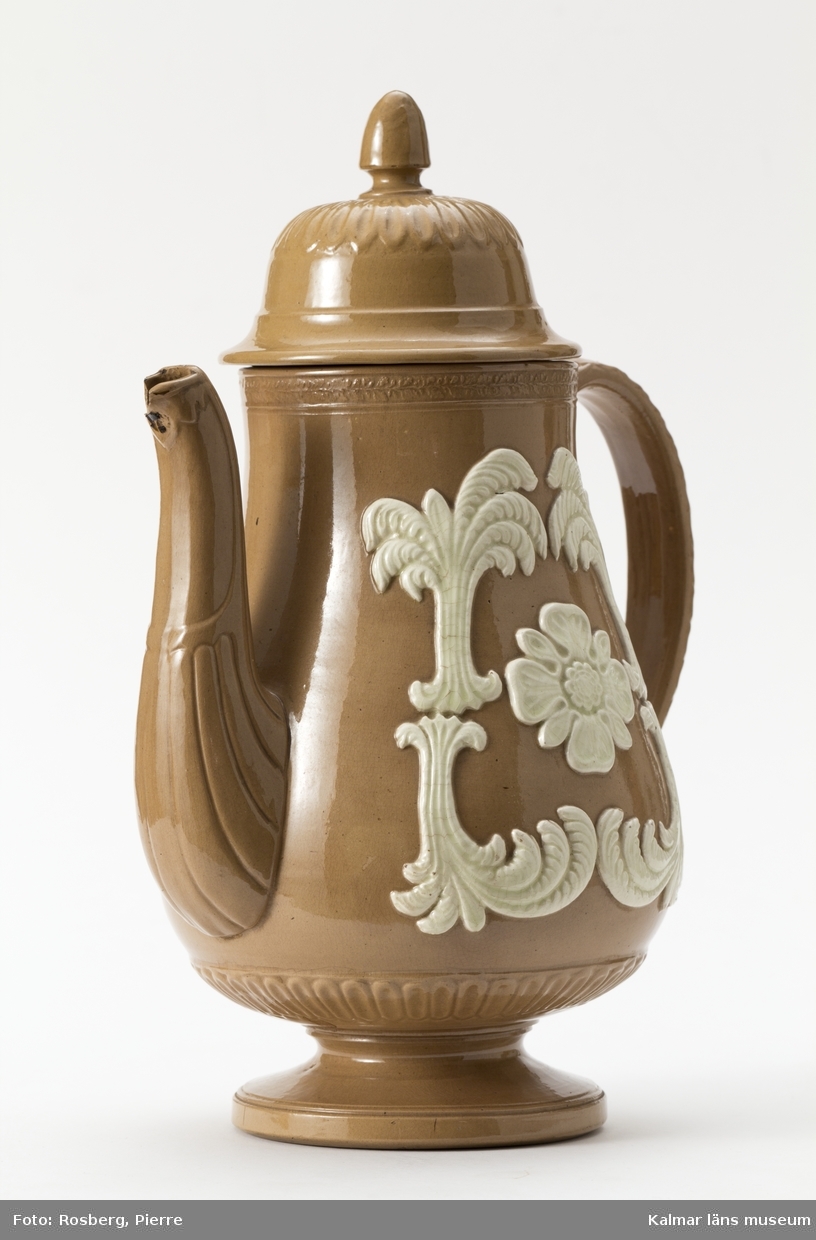 KLM 22815 Kaffekanna av lergods. Grågult gods, fajans med vit reliefdekor, palmkvistar och rosett. Stämpel i botten: T n F.
