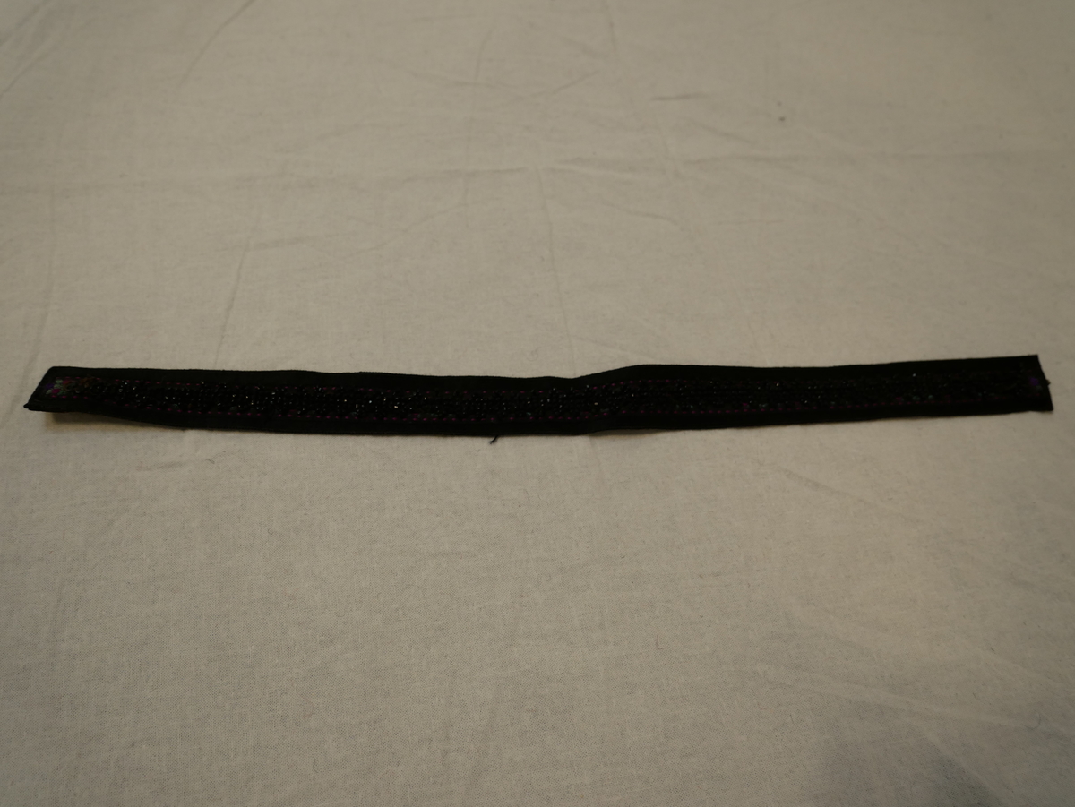 Svart silkeband med eit rosete silkeband sydd på langs midten. På silkebandet er set sydd fast eit band med svarte perler. Baksida fôra med brunt glansa lerret. Hekte i eine kortsida, krok i den andre.