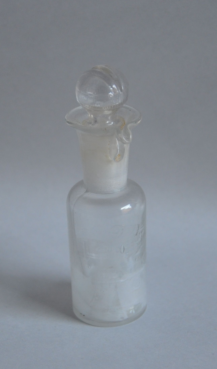Dråpeflaske av klart glass. Flasken har glasskork med spor slik at når korken vris sånn at sporet går mot den utbøyde munningen av flasken, kommer vesken dråpevis ut.